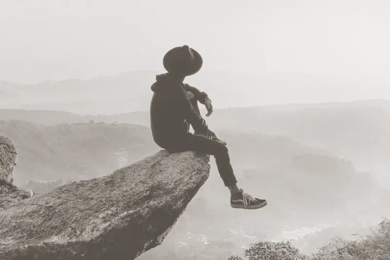 A person in outdoors hiking clothing and wide-brimmed hat, sitting balanced on the edge of a rock outcropping jutting into the frame