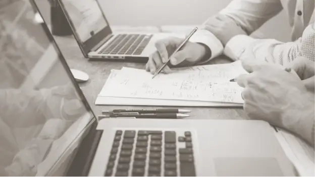 An office conference table, with laptops and hands taking notes